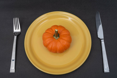 Little orange pumpkin on a yellow plate, fork and knife on black background