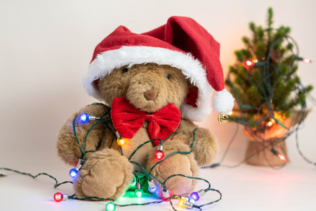 Cute teddy bear with red bow, Santa Claus hat and christmas lights on white background with...