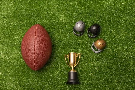 American football ball, little football helmets and golden trophy on green grass background