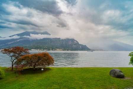 A serene lakeside landscape featuring calm water, lush green grass, and vibrant red trees in the foreground. Towering misty mountains surround the lake under a dramatic cloudy sky, with a small boat visible in the distance.の写真素材