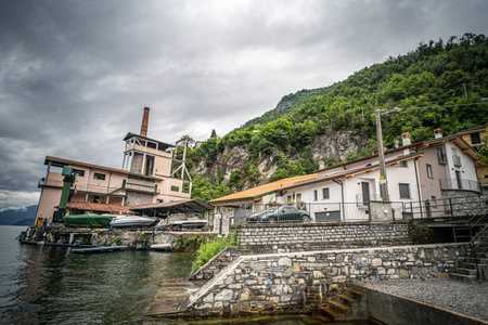 A lakeside village scene with rustic buildings nestled against a lush, green hillside under a cloudy sky. Boats are docked by the calm water, and stone pathways and retaining walls add a timeless charm to the setting.の写真素材