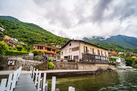 A picturesque lakeside village surrounded by lush green mountains under a cloudy sky. The scene features charming houses, including a lakeside restaurant, a wooden pier, and clear water reflecting the natural beauty of the setting.の写真素材
