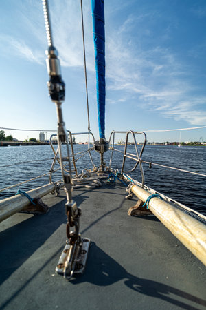 A close-up view of a sailboat deck featuring ropes, rigging, and a furled sail against calm waters and a clear blue sky. Coastal buildings and cranes are visible in the distance, adding a maritime touch.の写真素材
