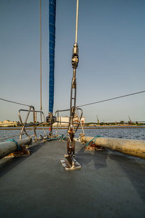 Close-up view of a sailboat focusing deck on rigging, ropes, and mast hardware. The calm water and harbor structures in the background highlight a peaceful maritime setting under clear skies.の写真素材