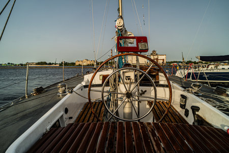 Close-up view of a sailboat cockpit featuring a classic wooden and metal steering wheel, ropes, and nautical equipment. Calm waters and docked boats are visible in the background under a clear sky.の写真素材