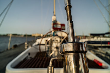 Detailed close-up of a sailboat's rigging and metal fittings with the helm and steering wheel visible in the blurred background. The scene captures a marina setting with calm waters and docked boats under a clear sky.の写真素材