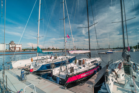 Sailboats docked at a marina pier with tall masts, colorful flags and calm waters reflecting a clear blue sky. Industrial buildings and cranes line the distant shore creating a serene waterfront sceneの写真素材