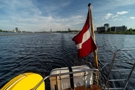 A red and white flag flutters at the stern of a sailboat cruising on a river, with a city skyline, bridge, and marina visible under a clear blue sky.の写真素材