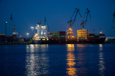 A cargo ship docked at twilight with cranes and stacked containers, reflecting lights on calm water.の写真素材