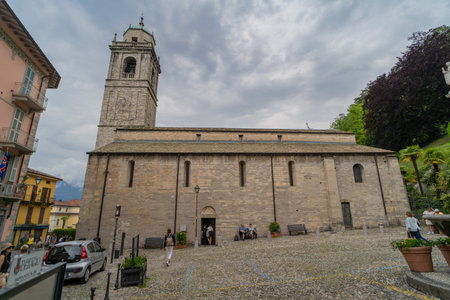 Historic stone church with a bell tower in a cobblestone plaza, surrounded by greenery, palm trees, and colorful buildings under a cloudy sky. Visitors are seen walking and sitting nearby.の写真素材