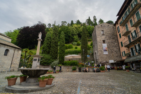 A picturesque European plaza featuring a stone fountain, historic buildings, and lush greenery on a hillside. Visitors are relaxing and strolling through the cobblestone square.の写真素材