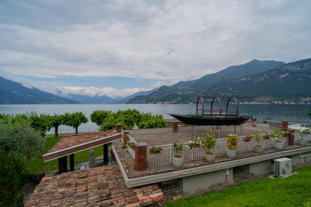 A scenic view of a lakeside terrace adorned with potted plants, overlooking mountains and calm waters under a cloudy sky. A decorative boat structureの写真素材