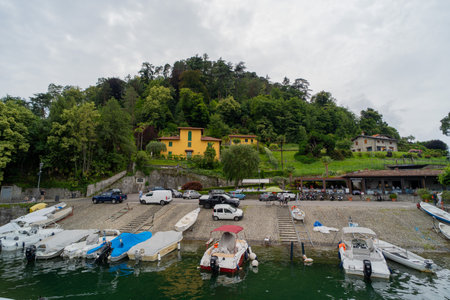 Scenic lakeside view with boats docked along the shore, colorful hillside houses, lush greenery, and a restaurant terrace overlooking the water.の写真素材