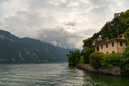 A serene lakeside villa surrounded by lush greenery and towering mountains under a cloudy sky, reflecting calm waters.の写真素材