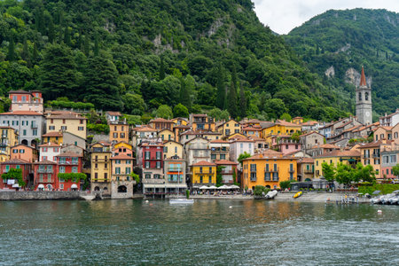 Colorful lakeside village with vibrant buildings, lush greenery, and a boat cruising on calm waters, surrounded by forested hills and a church tower in the background.の写真素材
