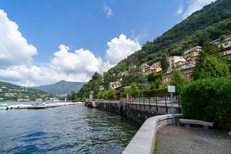 A scenic waterfront promenade along a lake, surrounded by lush green hills and elegant hillside villas under a bright blue sky with fluffy white clouds.の写真素材