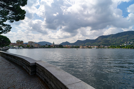 A serene lakeside promenade overlooking calm waters, with a picturesque town and lush green hills in the background under a partly cloudy sky.の写真素材