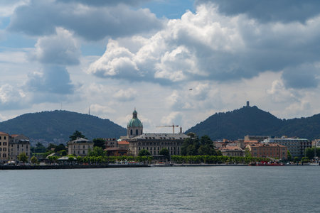 A scenic view of a lakeside city with historic architecture, including a domed cathedral, surrounded by lush green hills under a dramatic cloudy sky.の写真素材