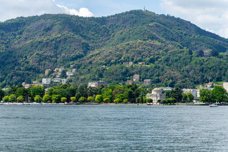 A serene lakeside view with lush green hills, elegant villas, and a classical building by the water, surrounded by trees and reflecting peaceful scenery under a bright sky.の写真素材