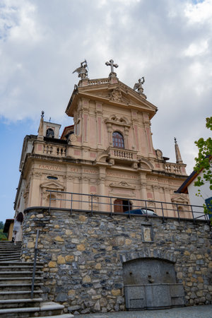 A historic Baroque-style church with ornate sculptures, intricate architectural details, and a stone wall foundation, set against a partly cloudy sky in a European town.の写真素材
