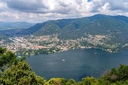 Panoramic view of a picturesque lakeside town surrounded by lush green mountains and calm waters, with boats sailing on the lake under a partly cloudy sky.の写真素材
