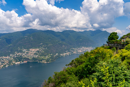 Scenic view of a tranquil lake surrounded by lush green hills and villages, with a clear blue sky and fluffy clouds overhead. A picturesque terrace overlooks the water.の写真素材