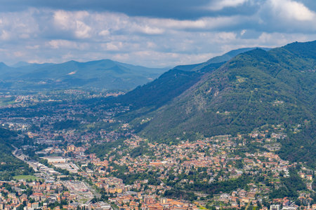 Aerial view of a town nestled at the foot of a green mountain, surrounded by rolling hills and distant peaks under a partly cloudy sky, showing casing urban and natural landscapes.の写真素材