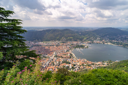 A panoramic aerial view of a city by a lake, surrounded by green hills. The scene features terracotta rooftops, a marina, and a cathedral under a partly cloudy sky.の写真素材