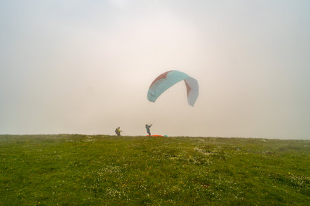 Paragliders preparing for takeoff on a foggy hillside meadow, with one parachute partially inflated, creating a scene of adventure and outdoor activity.の写真素材