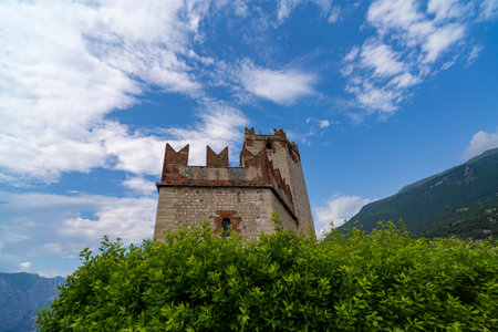 Historic stone castle tower with battlements rises above green foliage against a blue sky with scattered clouds and mountain slopes in the background.の写真素材