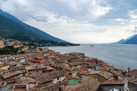 Charming lakeside village with terracotta rooftops, surrounded by green mountains and calm waters under a partly cloudy sky.の写真素材