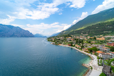 A picturesque lakeside town with colorful houses, a pebble beach, and lush green mountains rising along the shoreline under a partly cloudy sky.の写真素材