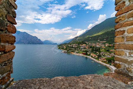 Scenic view of a lakeside town with colorful houses, pebble beaches, and green mountains, framed by ancient stone walls under a partly cloudy sky.の写真素材