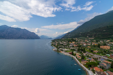 A picturesque lakeside town with colorful houses, lush greenery, and pebble beaches along a calm lake, surrounded by towering mountains under a partly cloudy sky.の写真素材