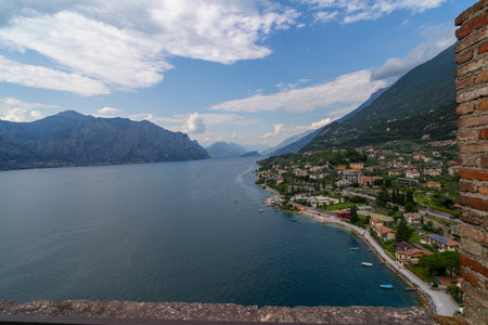Scenic view of a lakeside town with colorful houses, lush greenery, and pebble beaches, framed by mountains and calm waters under a partly cloudy sky.の写真素材