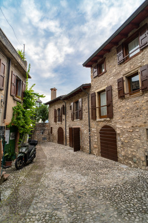Charming cobblestone alley lined with rustic stone houses featuring wooden shutters and arched doorways, set under a partly cloudy sky in a historic village.の写真素材