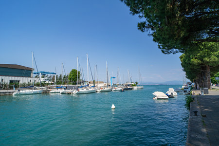 Scenic marina with sailboats and yachts docked along calm turquoise waters, surrounded by lush greenery and a clear blue sky. Peaceful promenade lined with trees.の写真素材