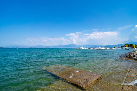 Crystal-clear waters of a serene lake with a small stone pier extending into the shallow shore. Sailboats and yachts docked at a distant marina under a bright blue sky.の写真素材