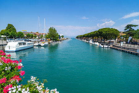 Scenic view of a canal lined with boats and yachts, surrounded by lush greenery, colorful flowers, and a tree-lined promenade under a clear blue sky.の写真素材