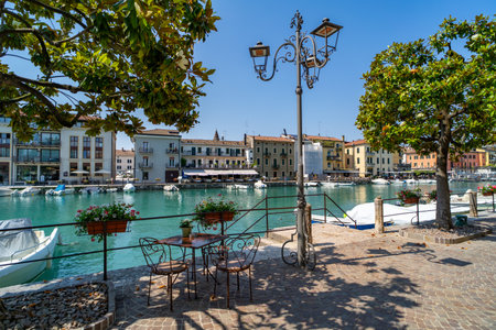 Scenic waterfront view of a European town with colorful buildings, boats docked along the canal, and a charming outdoor cafe table shaded by trees.の写真素材