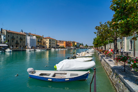 Picturesque canal scene in a European town with colorful buildings, moored boats, and a tree-lined promenade adorned with vibrant flowers. Calm waters reflect the architecture under a clear blue sky.の写真素材