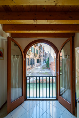 View through arched double doors overlooking a narrow Venetian canal and rustic buildings. The interior features wooden beams and tiled floors, blending charm with elegance.の写真素材