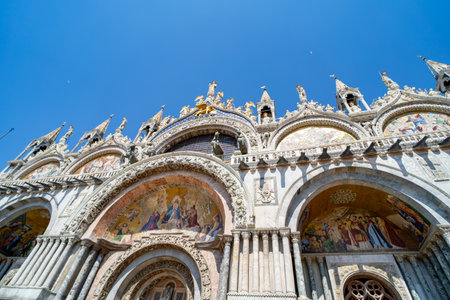 Facade of St. Mark's Basilica in Venice, Italy, showcasing intricate mosaics, arched entrances, and ornate sculptures against a clear blue sky.の写真素材