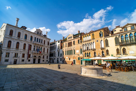 A charming Venetian square surrounded by historic buildings with ornate windows and colorful facades. Outdoor cafes and a stone well complete the lively atmosphere.の写真素材