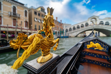 Close-up of a golden seahorse ornament on a Venetian gondola, with the iconic Rialto Bridge and colorful historic buildings in the background along the Grand Canal in Venice, Italy.の写真素材
