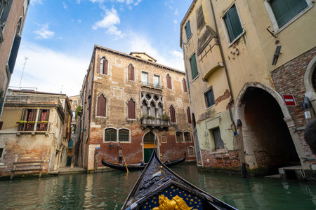 View from a gondola navigating a narrow canal in Venice, Italy, surrounded by historic buildings with Gothic windows and weathered facades under a partly cloudy sky.の写真素材