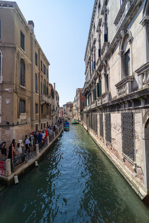A narrow canal in Venice lined with historic buildings and tourists walking along the edge, with small boats moored on the water under a clear sky.の写真素材