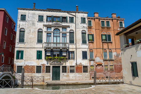 A row of historic Venetian buildings with colorful facades facing a canal, featuring arched windows, balconies, and green shutters under a clear blue sky.の写真素材