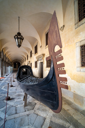 A historic black gondola with a detailed metal stern and decorative carvings, displayed in a Venetian arched hallway, symbolizing cultural heritage.の写真素材