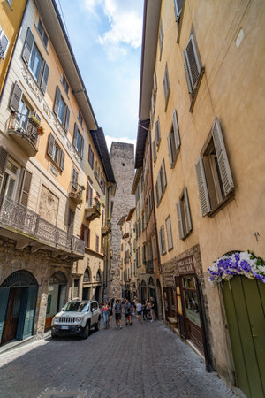 Picturesque view of a narrow cobblestone street in Bergamo, Italy, lined with colorful historic buildings and a medieval tower in the background.の写真素材